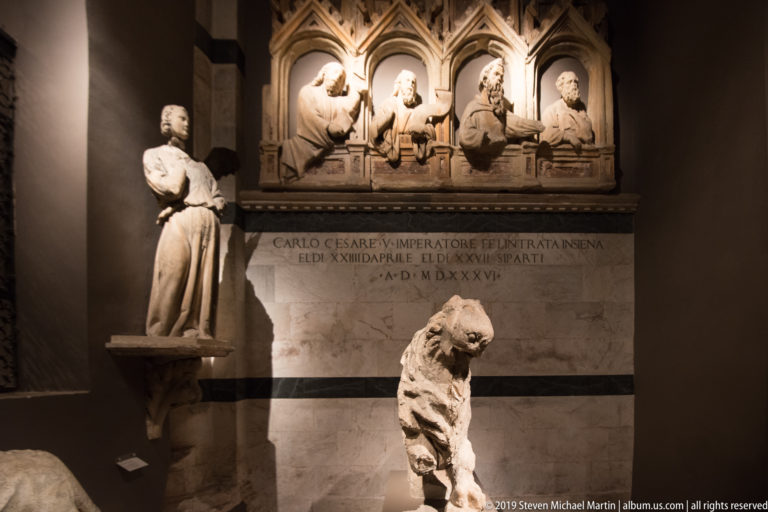 Marble wall monument with four busts beneath pointed canopies, a Latin inscription, standing figure, and weathered stone lion.