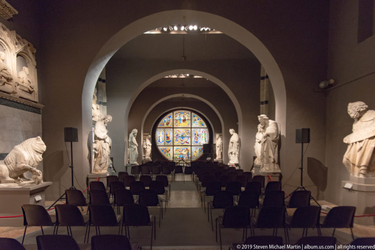 Long arched gallery lined with stone sculpture and chairs, terminating in a brightly illuminated circular stained-glass window.