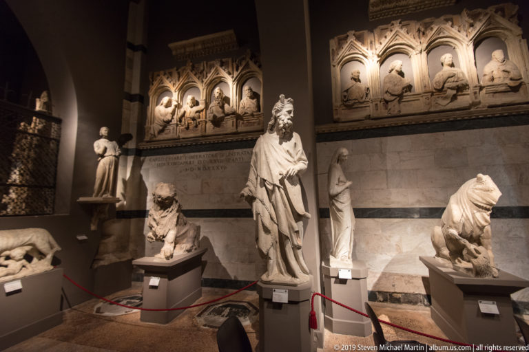 Stone figures and crouching lions displayed beneath a Gothic wall monument with pointed niches, busts, and an inscription.
