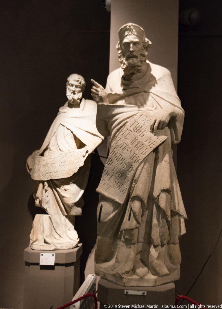 Two pale stone figures in a museum display, including a bearded figure holding a large inscribed tablet.