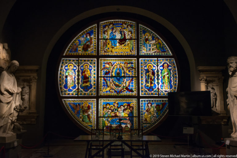Large circular stained-glass window glowing with multicolored sacred figures and narrative scenes in geometric compartments.