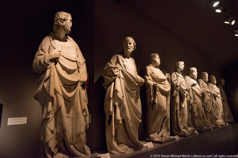Oblique row of monumental robed stone figures standing on a low museum platform under warm spotlights.