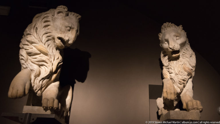 Large weathered stone lion with deeply carved mane and extended forelegs, dramatically lit against a dark museum wall.