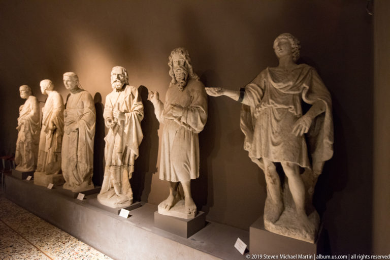 Row of standing stone statues on individual bases, including robed and bearded figures and a nearest figure in short classical-style dress.