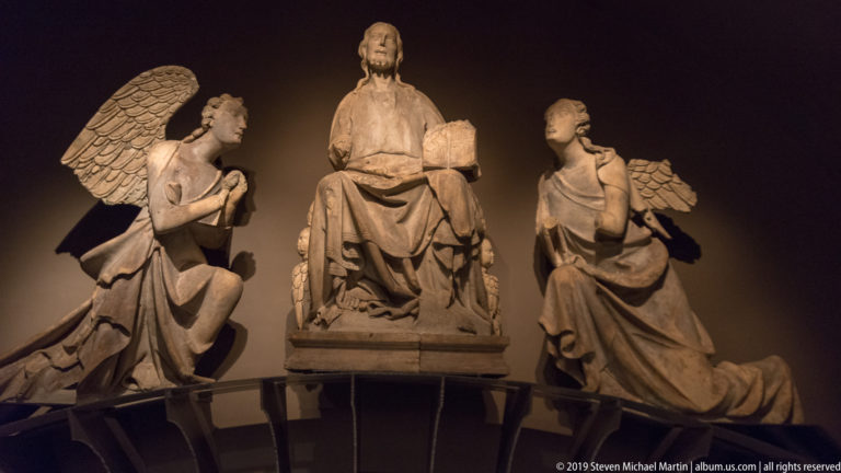 Stone group with a seated central figure flanked by two kneeling winged angels under warm museum lighting.