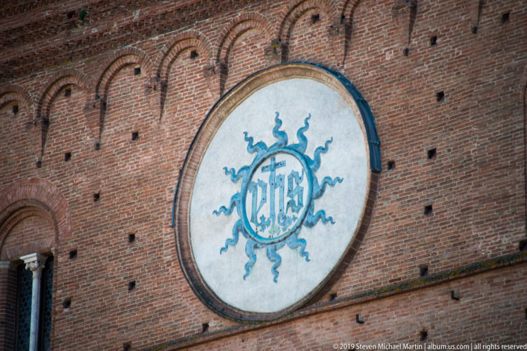 Large circular IHS Christogram with blue-green rays mounted on the red-brick façade of Palazzo Pubblico.