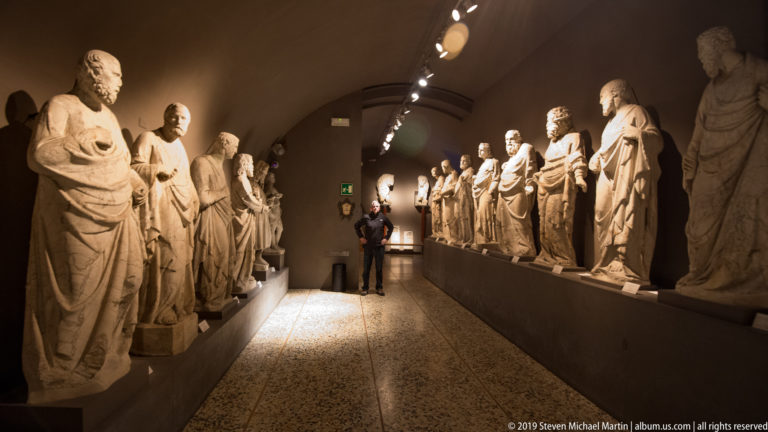 Long vaulted gallery with rows of standing stone statues on both sides and a visitor visible at the far end.