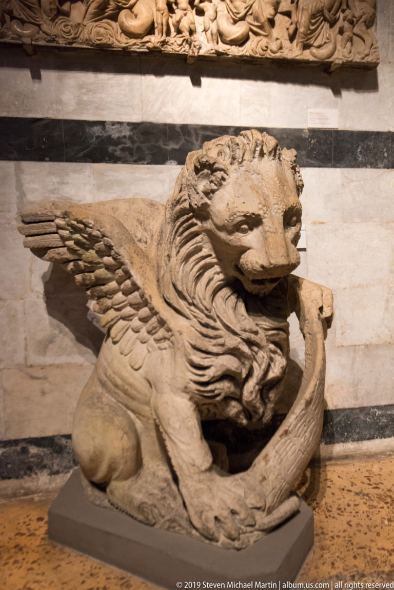 Carved stone winged lion crouching on a museum plinth, with folded feathered wings and a deeply textured mane.