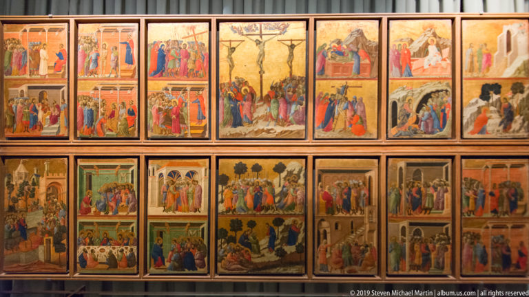 Wall display of many small framed religious narrative panels arranged in two rows, with figures, architecture, and landscape scenes.