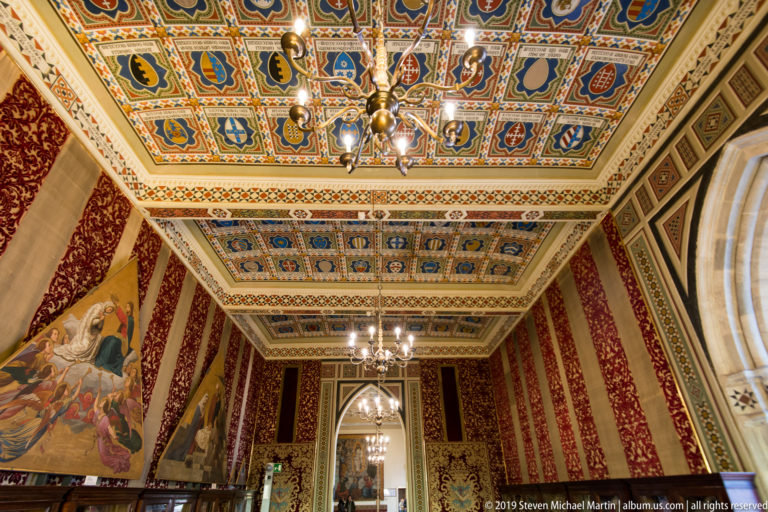 Historic interior with polychrome coffered ceiling, patterned red walls, mural paintings, chandeliers, and an arched doorway.