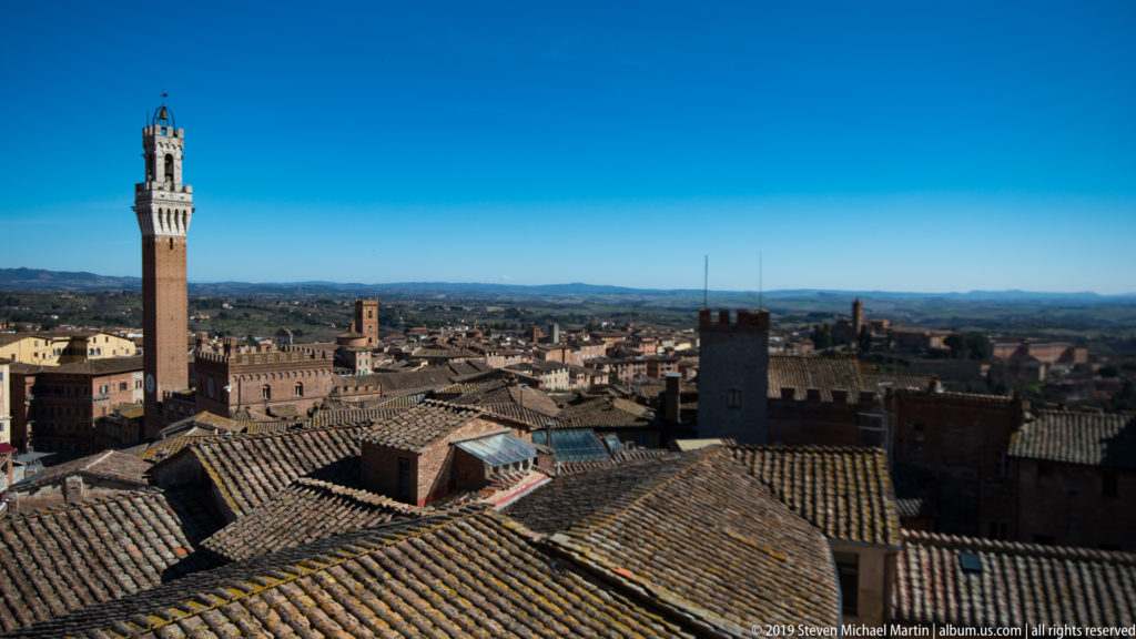 Torre del Mangia rising above layered terracotta roofs, chimneys, and masonry towers, with low Tuscan hills in the distance.