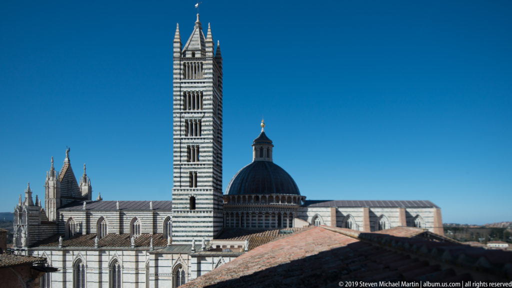 Striped Siena Cathedral campanile beside the dark dome and pinnacled roofline, seen above nearby terracotta roofs against blue sky.