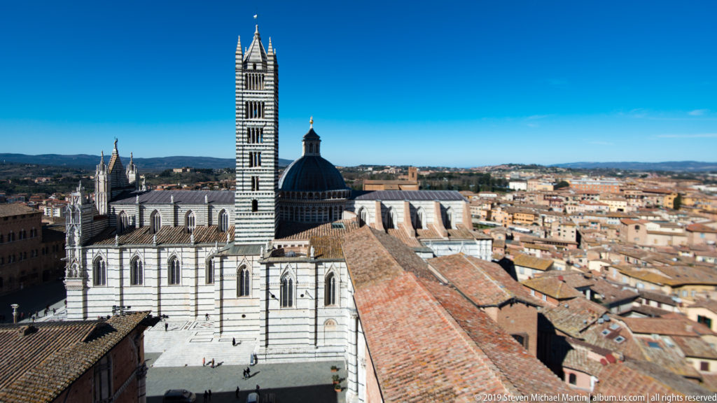 Elevated view of Siena Cathedral showing striped marble walls, tall striped campanile, dark dome, buttresses, and surrounding city.