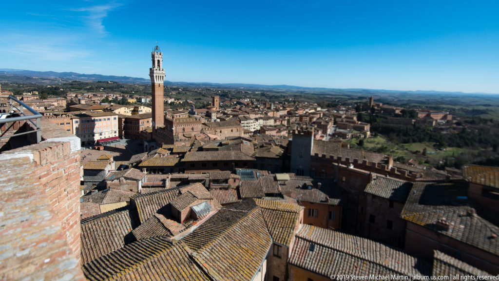 Elevated Siena roofscape with Torre del Mangia left of center and rolling countryside extending across the distant horizon.
