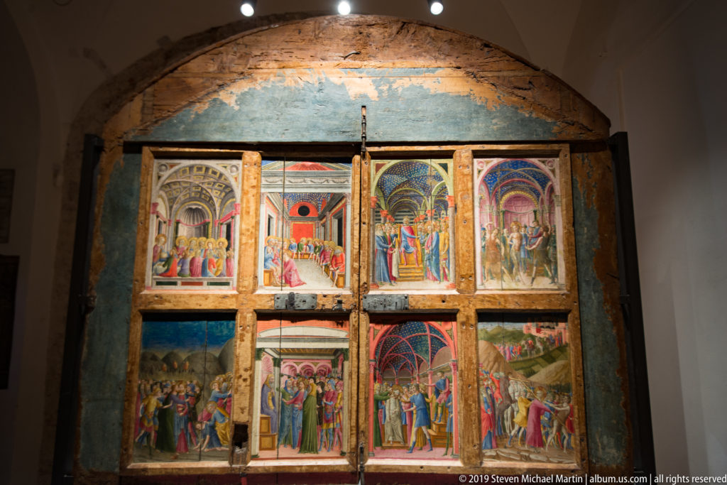 Large wooden altarpiece with multiple painted narrative panels in two tiers, displayed against a worn blue wall.