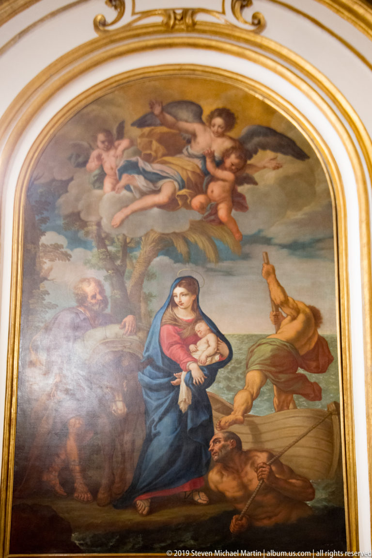 Arched painting of the Virgin in blue holding the Christ Child, surrounded by figures and angels beneath a gilded frame.