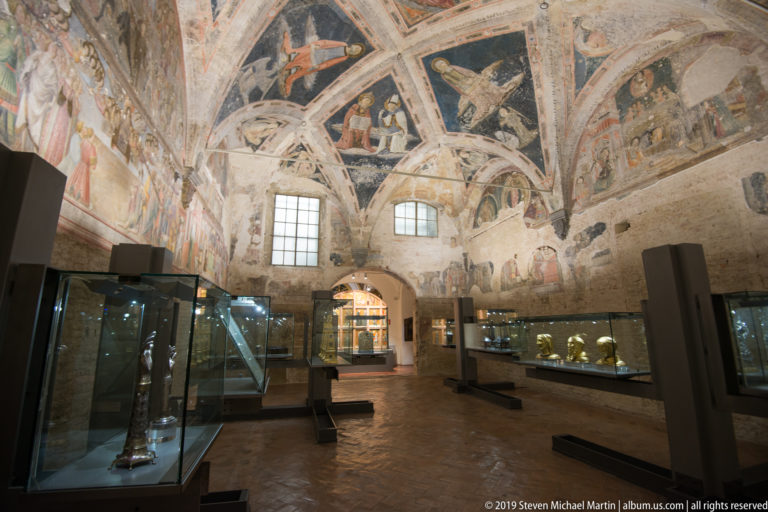 Historic vaulted museum room with fragmentary frescoes, two high windows, benches, and illuminated glass display cases.