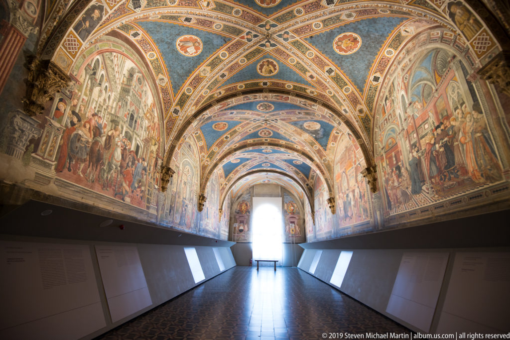 Symmetrical long view of a frescoed hall with blue rib vaults, murals on both walls, and a bright central doorway.