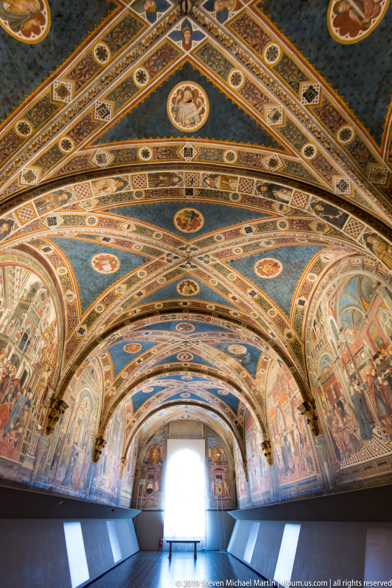 Long frescoed gallery with blue vaulted ceiling, narrative wall murals, museum panels, and a bright doorway at the far end.