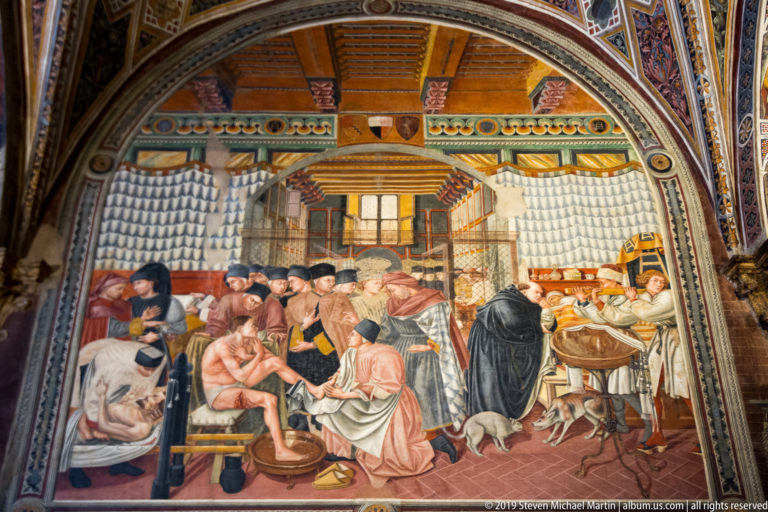 Fresco of caregivers tending patients in a ward lined with curtained beds, with attendants, food, and dogs nearby.