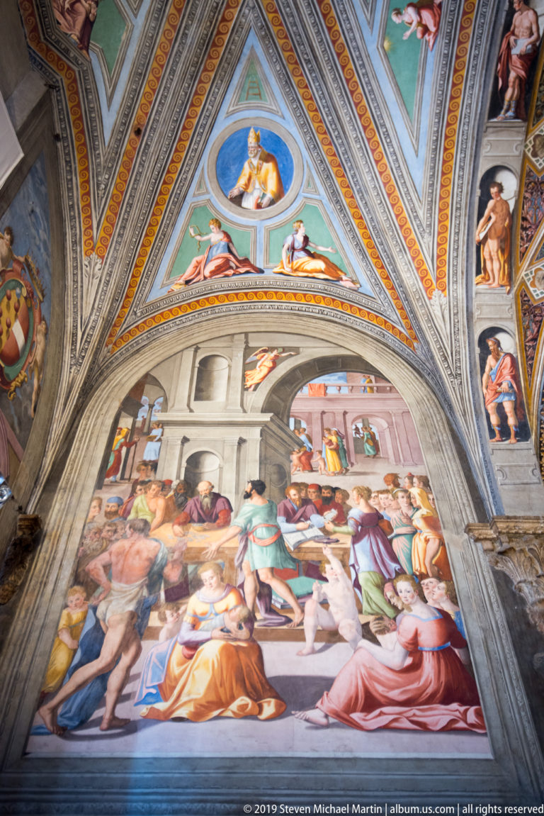 Fresco crowded with men and women beneath an arch, with painted triangular ceiling compartments and seated figures above.