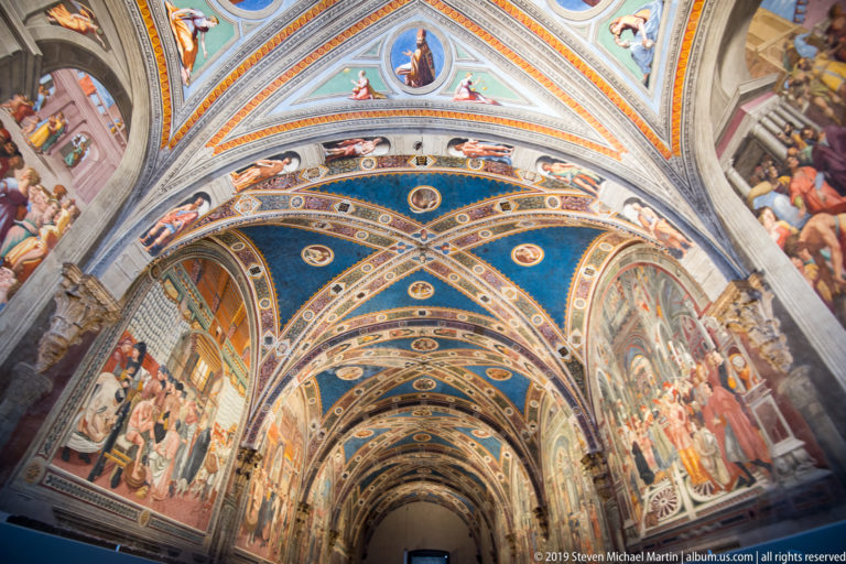 Long hall with blue frescoed rib vaults, circular medallions, and large narrative murals covering both walls.