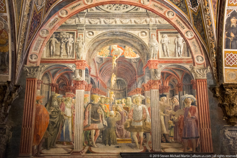 Fresco of mounted and standing figures gathered beneath a monumental arch with painted columns, reliefs, and a deep central passage.