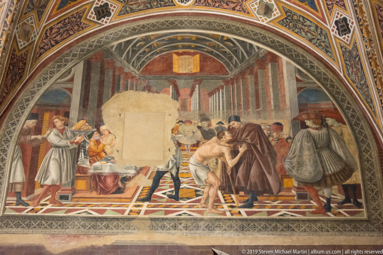Fresco of attendants caring for sick or injured people inside a long vaulted hospital ward with beds and numerous figures.