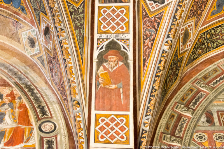 Painted bearded saint in a red robe holding a book, framed by geometric knotwork and ornate foliate borders.