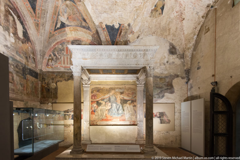 Stone shrine with fluted columns and carved entablature framing a religious fresco in a room of fragmentary wall paintings.