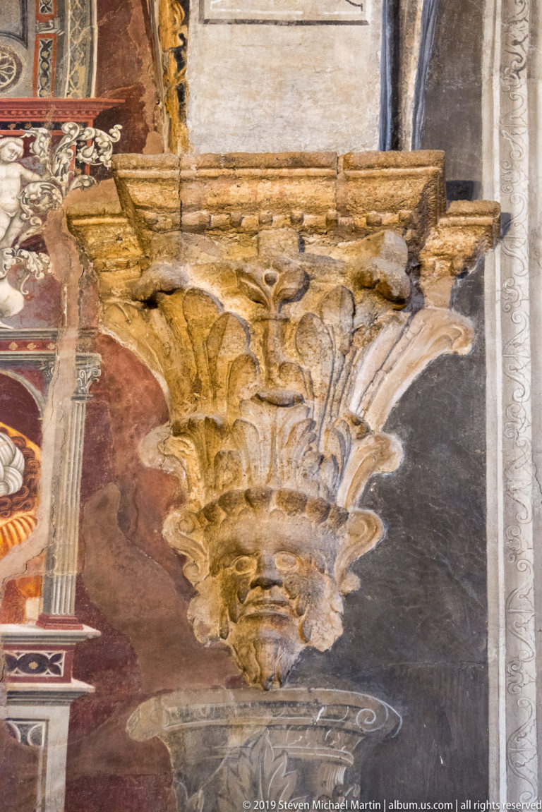 Gilded carved capital with layered leaves and scrolls projecting beside painted columns and ornamental wall decoration.