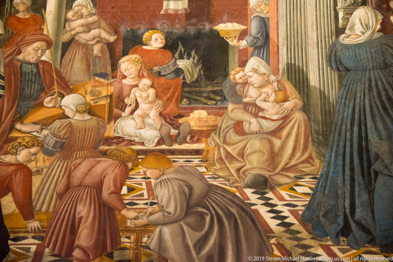 Fresco with women holding infants, attendants, visitors, and patterned floor inside a richly decorated architectural interior.