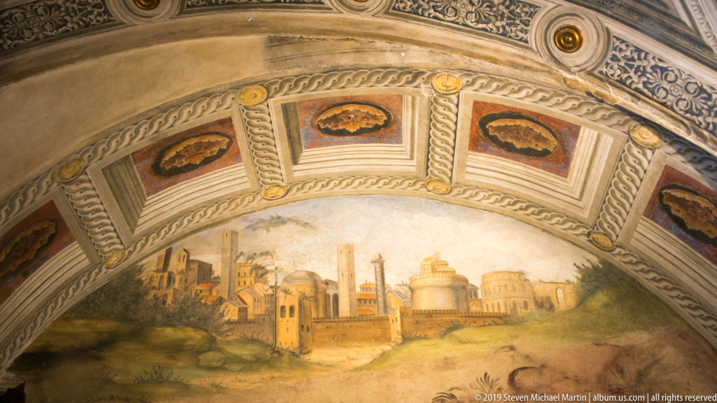 Semicircular painted cityscape of towers and domes beneath an ornate ribbed vault with patterned coffers.