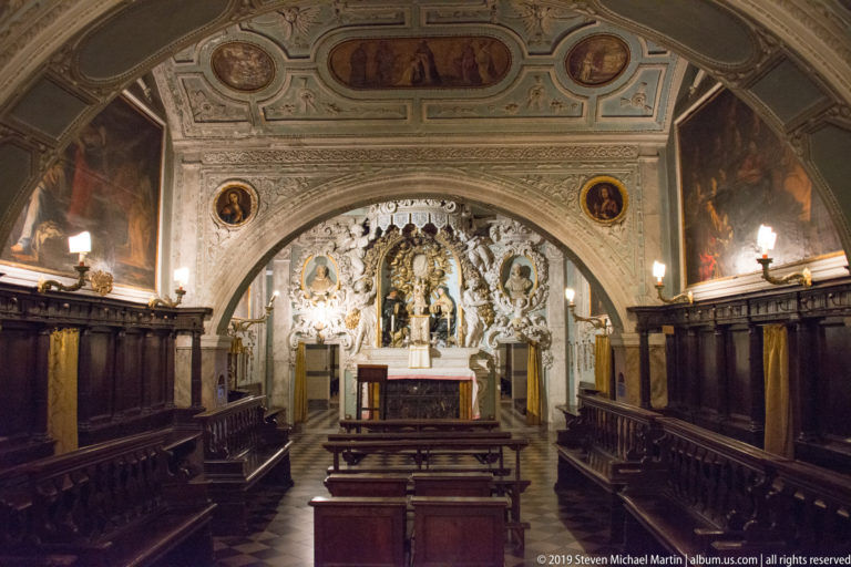 Axial chapel view with wooden stalls and benches leading beneath a large arch to an ornate white-and-gold altar.
