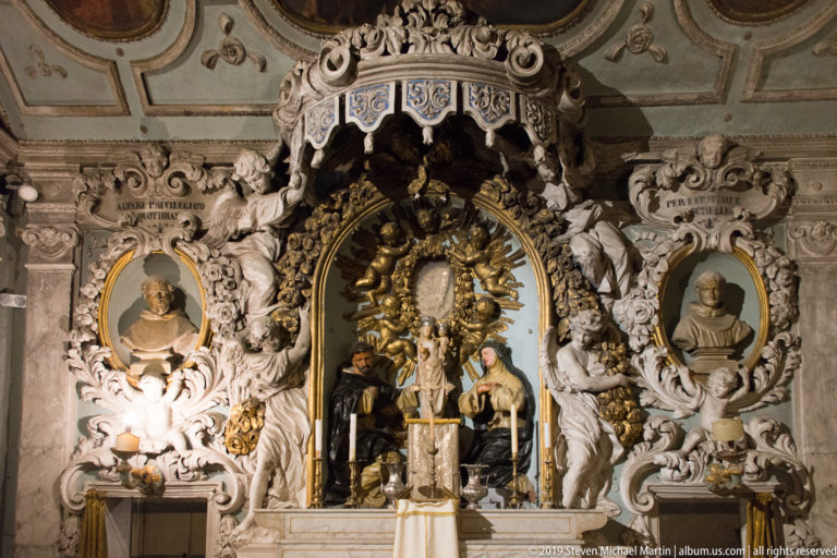 Ornate white-and-gold altar with central sacred image, sculpted figures, portrait busts, and a canopy-like crown.