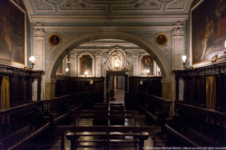 Long chapel interior with dark wooden choir stalls, pale stucco arches, painted ceiling, and devotional decoration at the far end.