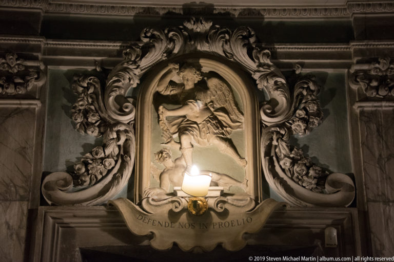 Ornate devotional shrine with central sacred relief, portrait medallions, sculpted figures, gilding, and the inscription “DEFENDE NOS IN PROELIO.”