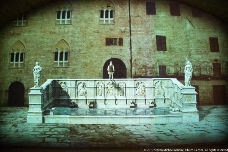 Historic view of a rectangular relief-covered fountain with corner statues before a large stone building.