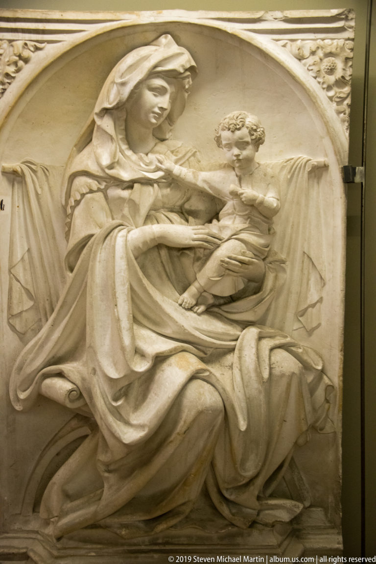 Arched stone relief of the seated Virgin supporting the Christ Child across her lap with one arm extended.