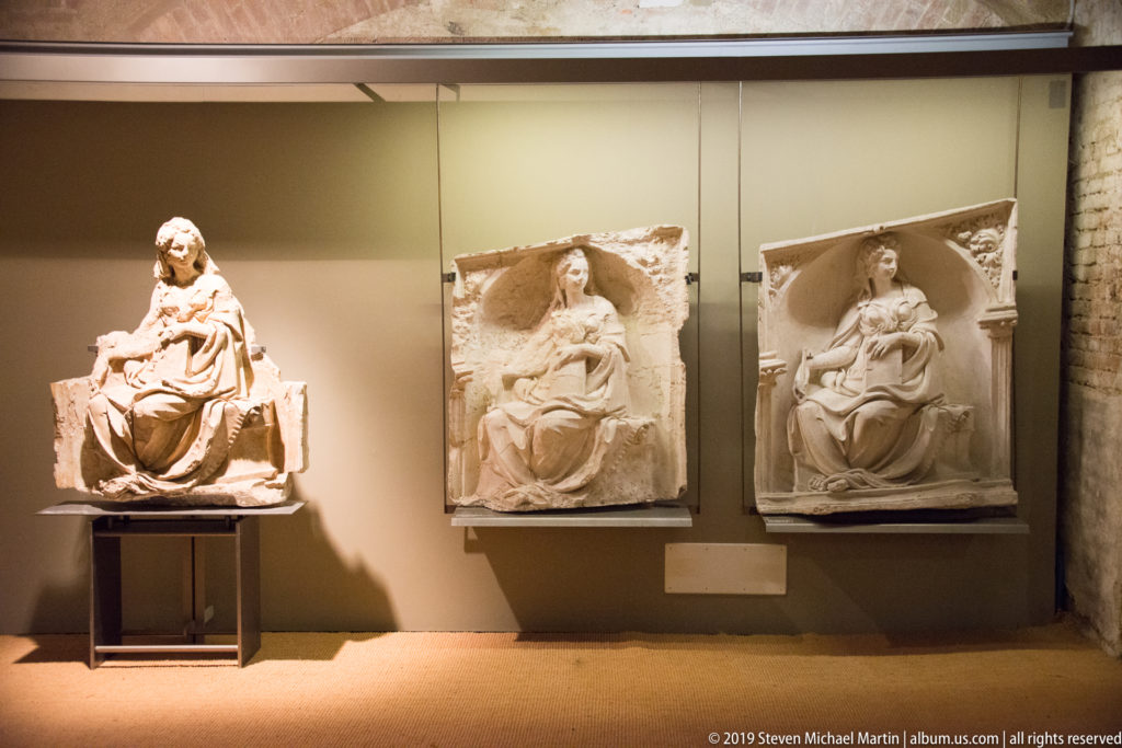 Three seated stone devotional sculptures displayed together, one freestanding and two mounted as relief panels.
