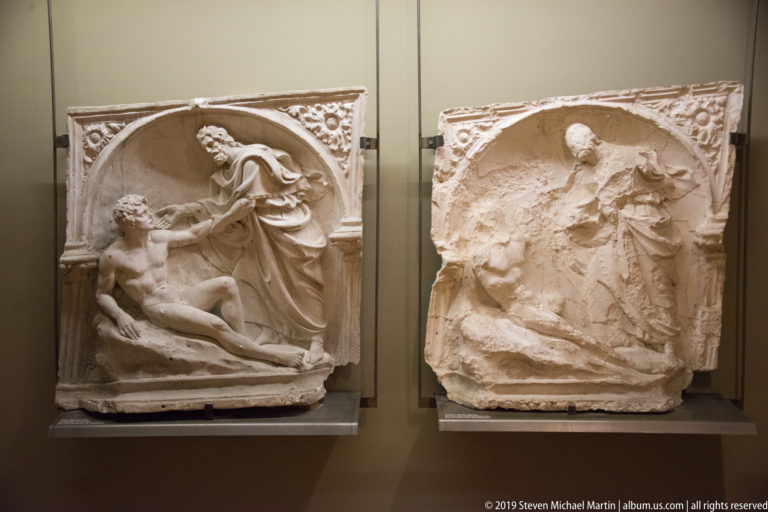 Two arched stone reliefs with crowded biblical-style figures, including a reclining nude man and robed woman at left.