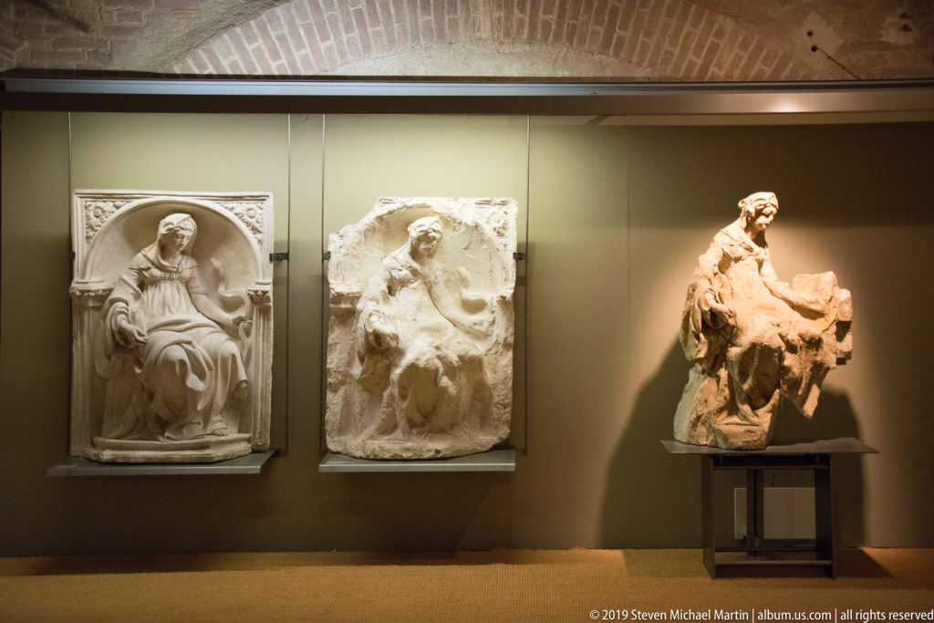 Three seated stone devotional sculptures displayed together, two mounted on the wall and one on a stand.