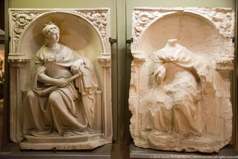 Two arched stone reliefs of seated women, one largely intact and one heavily damaged and headless.