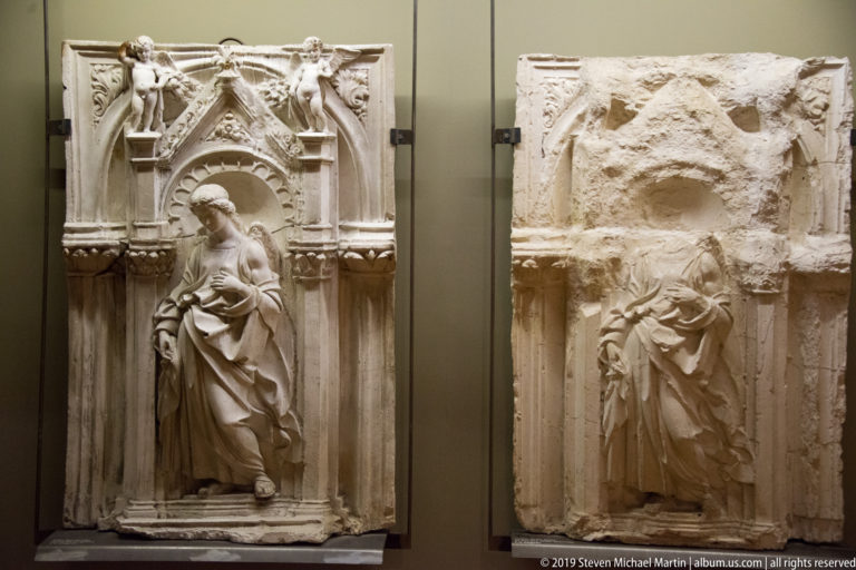 Two tall stone reliefs of standing robed figures beneath Gothic niches, one intact and one heavily damaged.
