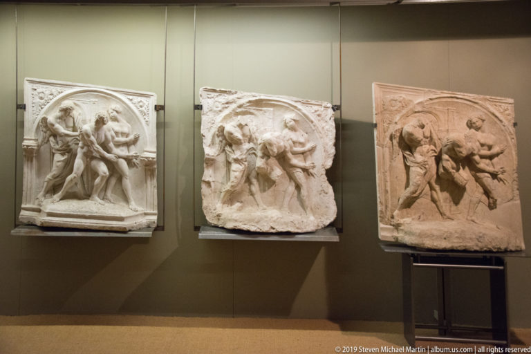 Three museum-mounted stone reliefs with crowded groups of carved human figures in shallow architectural frames.