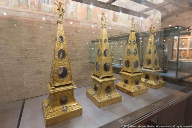 Four tall gilded pyramidal objects with stepped bases, finials, and dark oval insets displayed together in a glass case.