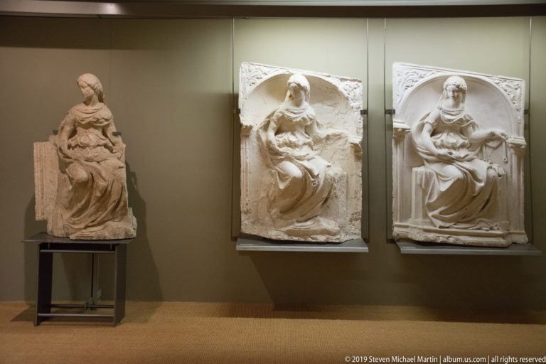 Museum wall with a seated stone Madonna sculpture and two arched reliefs of seated female figures.