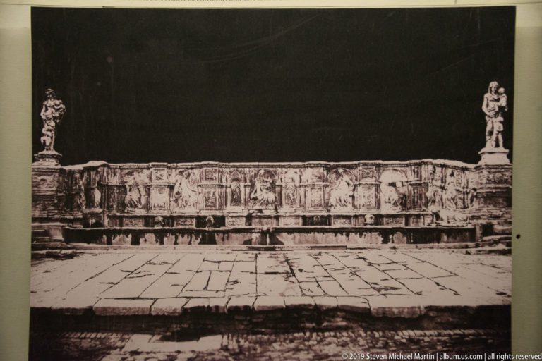 Historic black-and-white view of a long carved fountain with relief panels and standing statues at its ends.