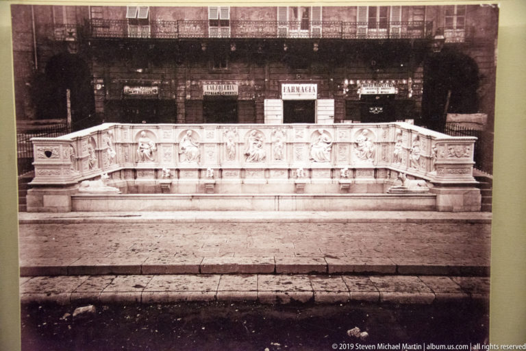 Historic black-and-white photograph of a long rectangular marble fountain with carved relief panels in a paved square.