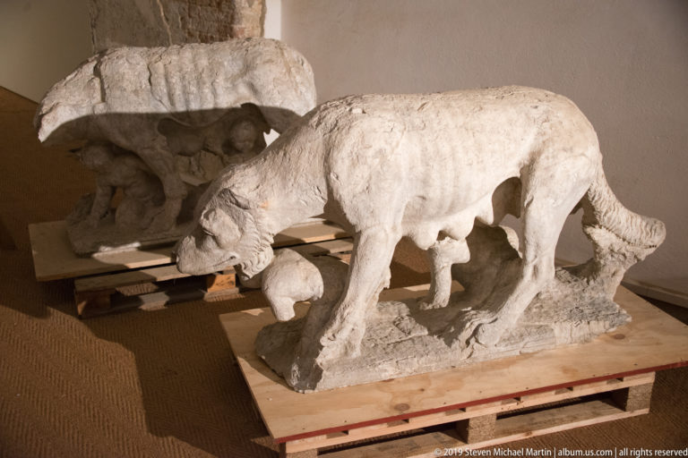 Weathered marble animal sculpture standing over a small nursing figure on a wooden pallet beside stone fragments.